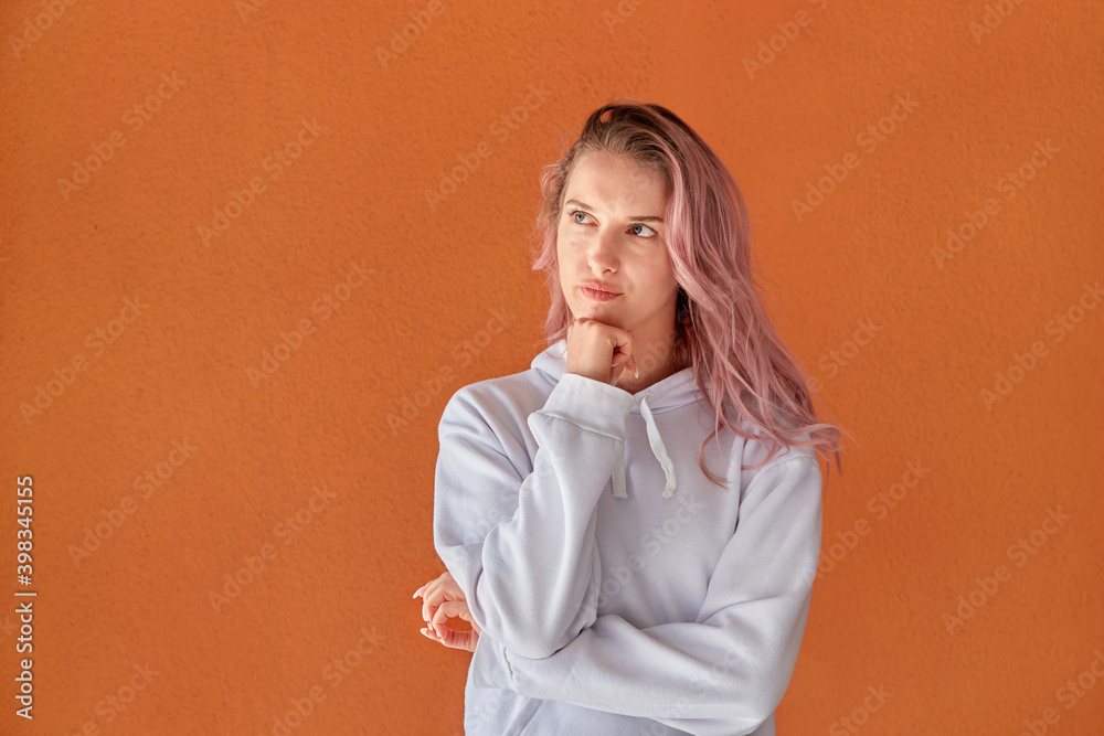 Cute woman with pink hair thinks and looks away. The concept of choice. Orange background.