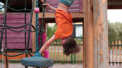Boy in an orange T-shirt hangs upside down in the exterior playground. Training endurance, agility and coordination of children in their free time. Healthy lifestyle. Summer. Outdoors.