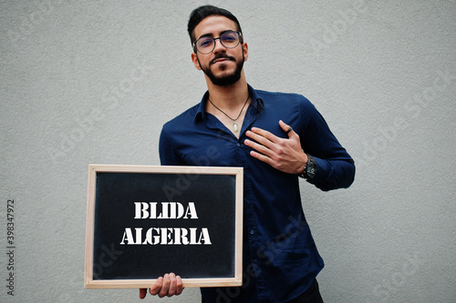 Arab man wear blue shirt and eyeglasses hold board with Blida Algeria inscription. Largest cities in islamic world concept. photo