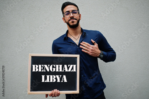 Arab man wear blue shirt and eyeglasses hold board with Benghazi Libya inscription. Largest cities in islamic world concept. photo