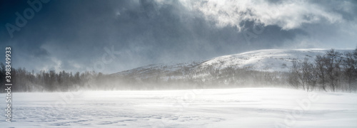 Panoramic view on mountain snow drift in Swedish Lapland. Sunny day, high mountain range with wild arctic birch forest at the lower level. High speed clouds and drifting snow. Scandinavian mountains.
