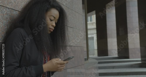 Side portrait of the young gorgeous african girl enjoying music in earphones, dancing and text ing via the mobile phones while leaning on the wall outdoor.
