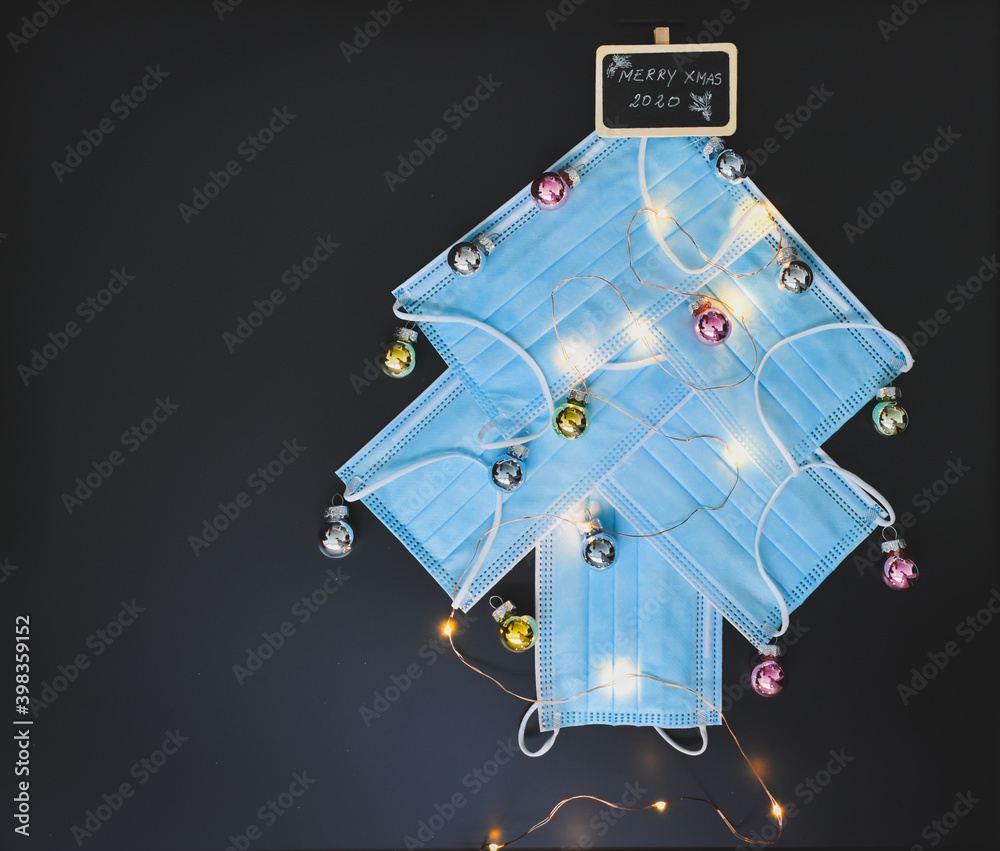 View of a Christmas tree made of blue surgical masks decorated with colorful Christmas baubles and Christmas lights against black chalkboar background. Christmas during pandemic or medical background.