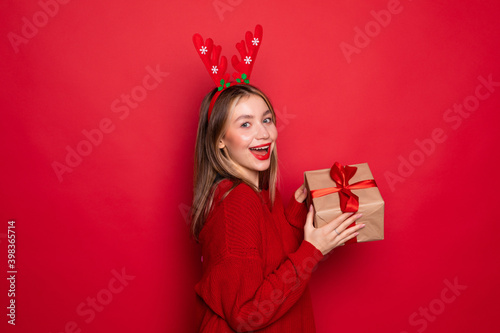 Amazed young Santa girl in fun decorative deer horns on head pointing index finger on red boxes with gifts presents on red background. Happy New Year 2019 celebration holiday party concept
