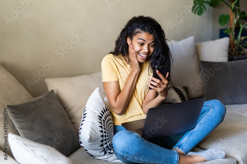 Surprised woman using smart phone and laptop sitting on a couch in the living room at home