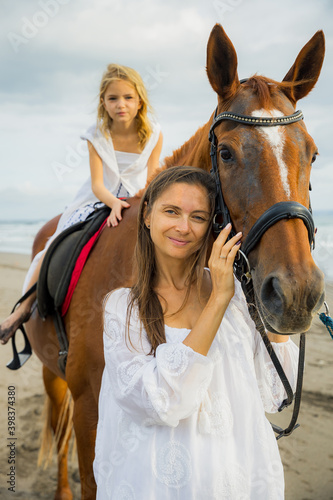 Horse riding on the beach. Cute little girl on a horse. Her mom stroking a horse. Portrait of woman and brown horse. Love to animals. Mother and daughter spending time together. Selected focus.