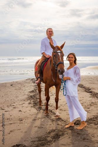Horse riding on the beach. Handsome man on a brown horse. Woman standing near by and stroking a horse. Love to animals. Husband and wife spending time together. Selected focus.