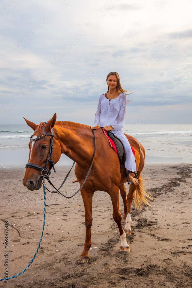 Beautiful caucasian woman riding horse on the beach. Female wearing white clothes. Copy space. Sunset time on the beach. Outdoor activities. Beautiful caucasian woman riding horse on the beach. Female wearing white clothes. Copy space. Sunset time on the beach. Outdoor activities.