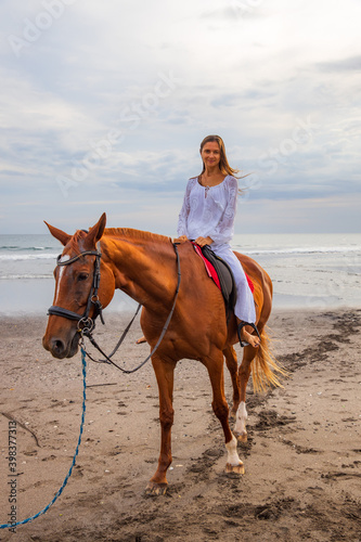 Beautiful caucasian woman riding horse on the beach. Female wearing white clothes. Copy space. Sunset time on the beach. Outdoor activities.