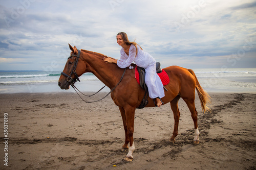 Horsewoman lays down on the withers of the horse. Caucasian woman in white dress riding horse on the beach. Copy space. Sunset time on the beach. Outdoor activities.