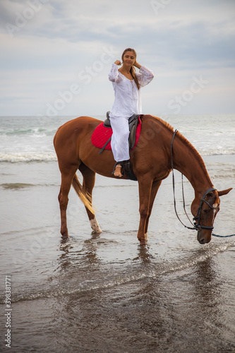 Beautiful caucasian woman riding horse by the sea. Female wearing white clothes. Copy space. Sunset time on the beach. Outdoor activities.