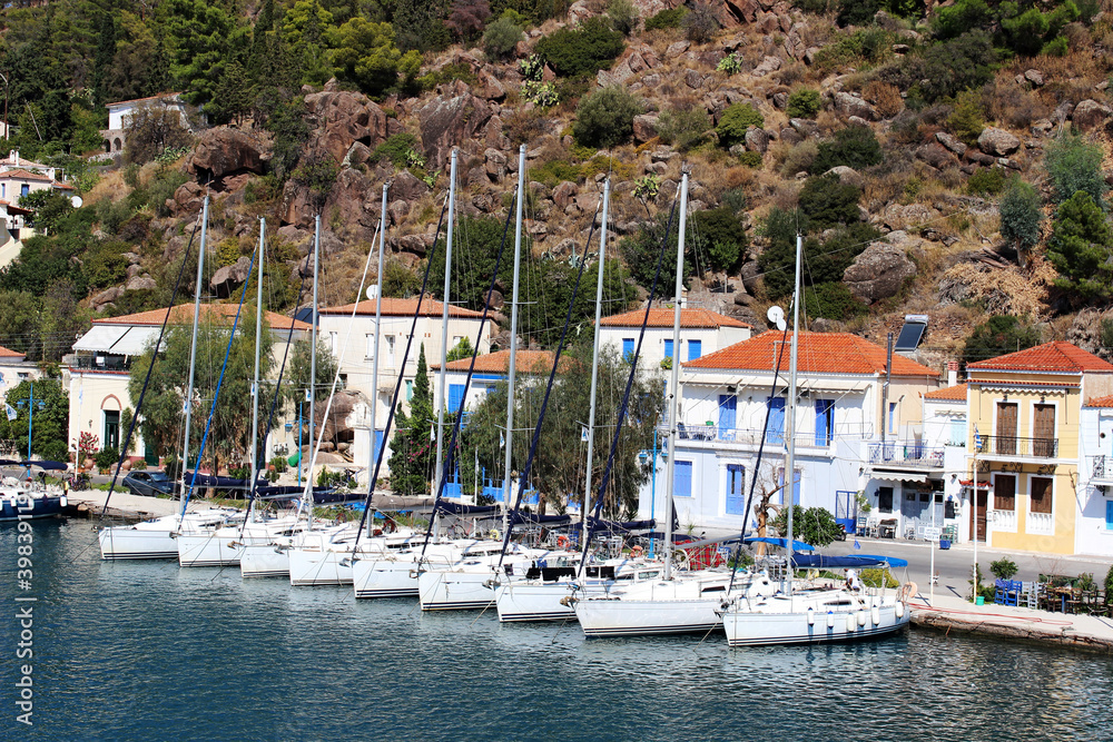 An old Mediterranean town with white houses and red roofs on a mountainside. There are white boats in the port. The bright sky is reflected in the blue sea.