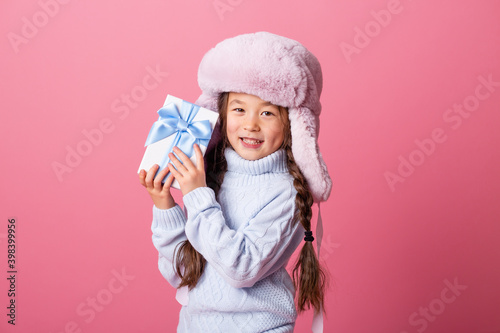 Cute little Asian girl in a winter hat and sweater holds a gift box. Christmas concept, text space