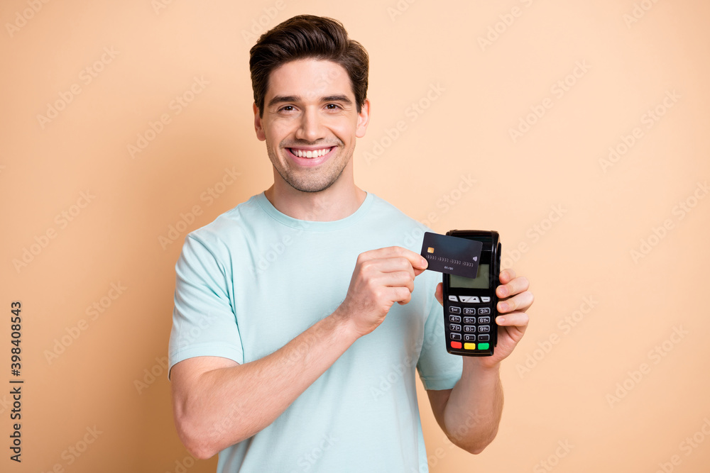 Close-up portrait of nice cheerful guy holding in hand terminal using bank card finance transaction isolated over beige pastel color background - obrazy, fototapety, plakaty 