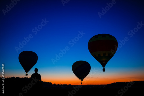Three Amazing Colorful Air Balloons Levitating Over the Ground Outdoors With A human Silhouette Against Blue Skies At Twilight.