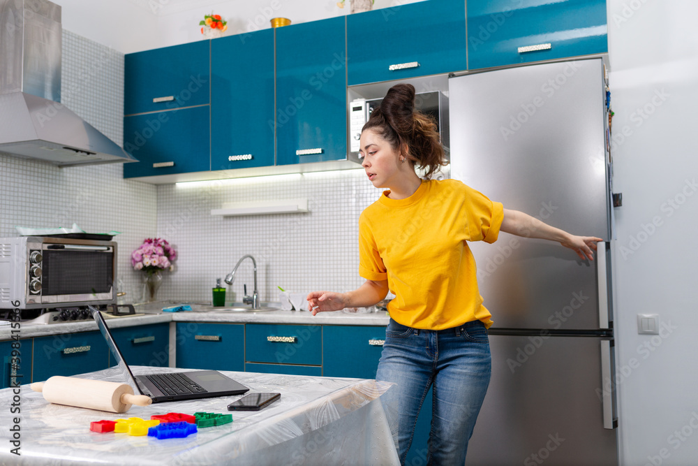 A young beautiful woman opens the refrigerator and looks carefully at her laptop. Indoor. The concept of cooking at home using online recipes and video communication