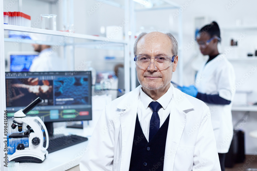 Senior scientist sitting at his workplace during medical experiments. Elderly scientist wearing lab coat working to develop a new medical vacine with african assistant in the background. Senior scientist sitting at his workplace during medical experiments. Elderly scientist wearing lab coat working to develop a new medical vacine with african assistant in the background.