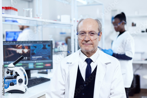 Senior scientist sitting at his workplace during medical experiments. Elderly scientist wearing lab coat working to develop a new medical vacine with african assistant in the background.