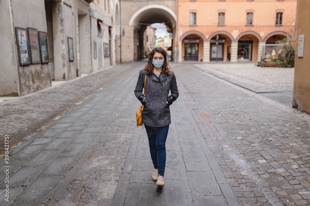 Close-up portrait caucasian smiling young woman walking in a city center during cold winter season. Lady wearing a protective face mask. Pandemic, new normal concept.