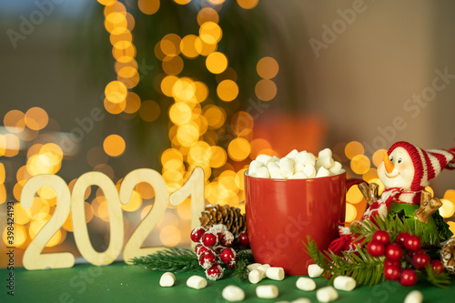 A red mug with cocoa and marshmallows next to a snowman. Christmas decor with a bright garland. Selective focus, shallow depth of field, background. 2021.