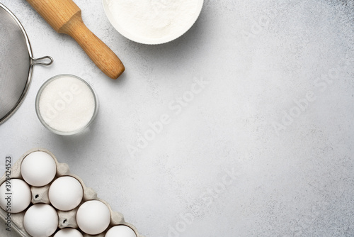 Flat lay with baking ingredients for pastry. Wheat flour, sugar, sieve, rolling pin and white eggs on the grey background. Top view, copy space. 