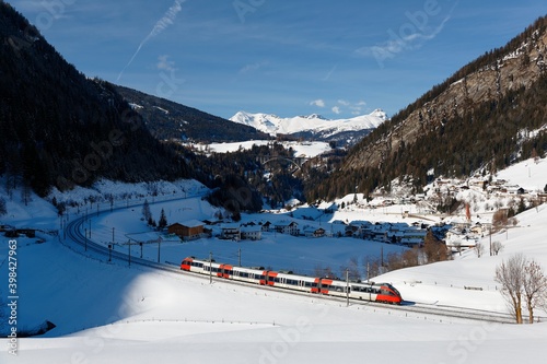A train traveling through the railway curve in a valley covered by heavy snow and St. Jodok village in Brenner Pass lying on the hillside of Alpine mountains on a sunny winter day, in Tyrol, Austria
