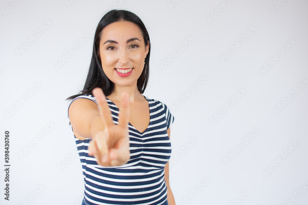 Portrait of gorgeous brunette woman in striped t-shirt looking at camera with smile and showing peace sign with fingers isolated over white background Portrait of gorgeous brunette woman in striped t-shirt looking at camera with smile and showing peace sign with fingers isolated over white background