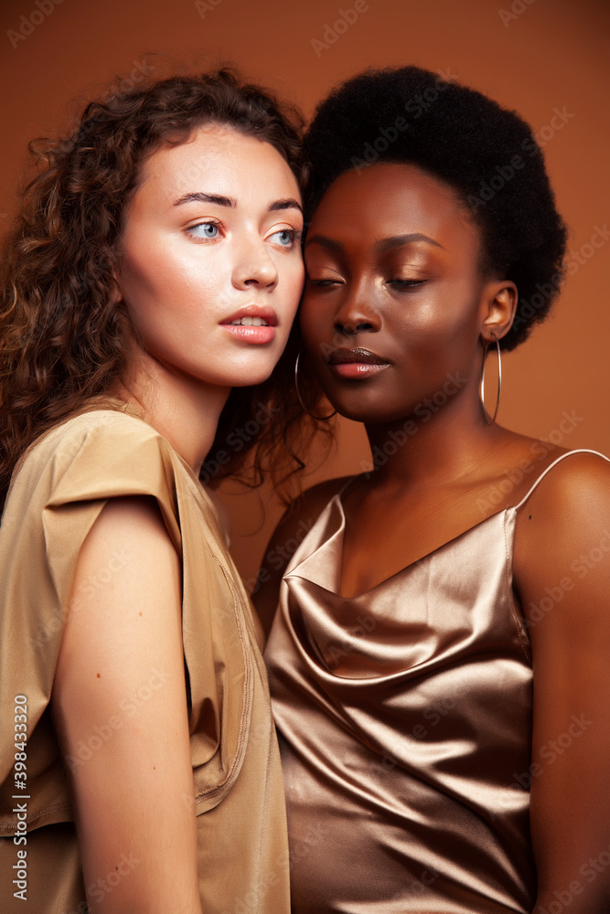 two pretty girls african and caucasian blond posing cheerful together on brown background, ethnicity diverse lifestyle people concept