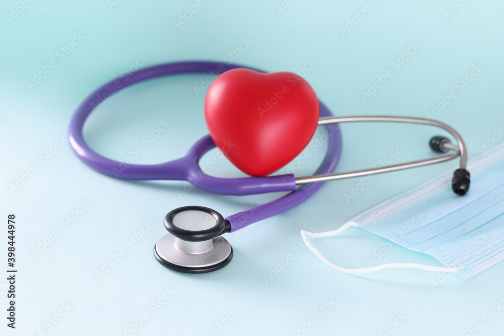 Lilac stethoscope and red toy heart with protective medical mask closeup Lilac stethoscope and red toy heart with protective medical mask closeup