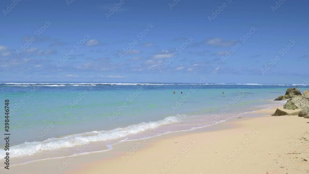 Tropical beach with small waves, vibrant blue water, yellow sand with stones and people silhouettes are swimming in background Tropical beach with small waves, vibrant blue water, yellow sand with stones and people silhouettes are swimming in background