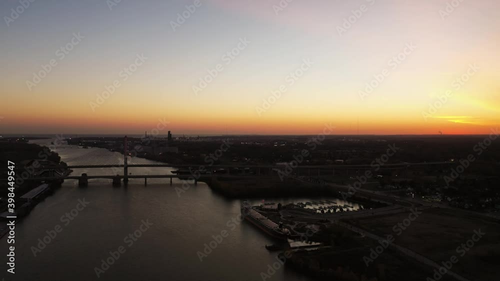 Aerial flying above the Maumee River in Toledo Ohio with bridges, a docked ship and industrial buildings below and the Lake Erie coastline at the horizon beyond below a orange and pink sunrise sky. Aerial flying above the Maumee River in Toledo Ohio with bridges, a docked ship and industrial buildings below and the Lake Erie coastline at the horizon beyond below a orange and pink sunrise sky.