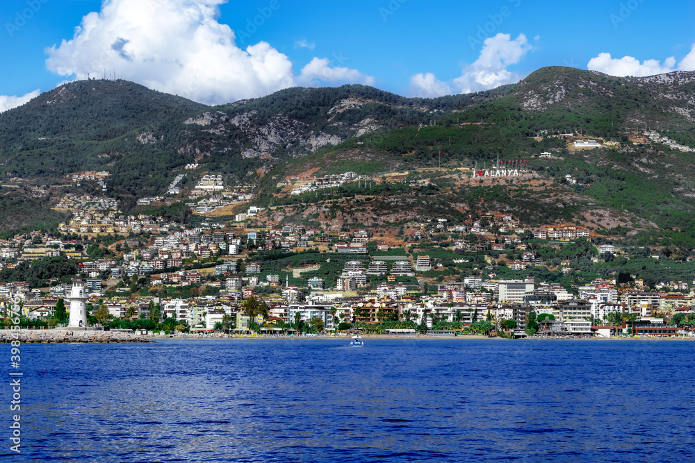 Alanya (Turkey) coastline panorama - view from Mediterranean Sea. I love Alanya sign on the hillside and a lighthouse in the water against the background of a modern tourist town among the mountains