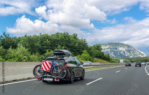 Black car with roof luggage box and trunk bike rack driving on highway. Beautiful mountain landscape background