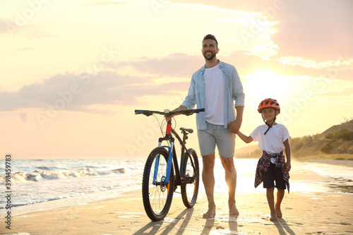 Happy father and son with bicycle on sandy beach near sea at sunset