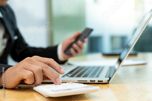 Business man using mobile smart phone, busy working on laptop computer browsing internet or checking internet application on smartphone with digital tablet and business data on office desk, close up