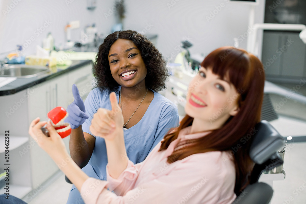 Obraz premium Dentist and patient in clinic. Oral care, orthodontics. Smiling happy African woman dentist and her female patient holding together teeth model, looking at camera with thumbs up. Focus on dentist