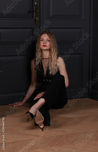 A pretty blonde with curly hair, in black festive clothes, sits on the floor, in the room, by the door and looks sad. Cheating, divorce, stylish pose, disappointment, reason to think, corporate,