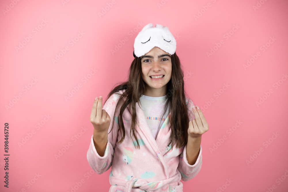 Pretty girl wearing pajamas and sleep mask over pink background doing money gesture with hands, asking for salary payment, millionaire business Pretty girl wearing pajamas and sleep mask over pink background doing money gesture with hands, asking for salary payment, millionaire business