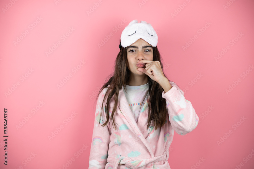 Pretty girl wearing pajamas and sleep mask over pink background mouth and lips shut as zip with fingers. Secret Pretty girl wearing pajamas and sleep mask over pink background mouth and lips shut as zip with fingers. Secret