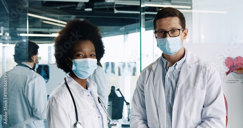 Close up portrait of happy African American female doctor standing with Caucasian male colleague and looking at camera in clinic office in good mood during coronavirus pandemic. Health concept