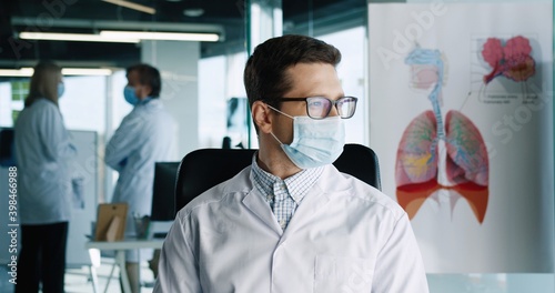 Close up portrait of handsome young Caucasian adult male doctor wearing medical mask sitting in clinic at workplace looking at camera and smiling. Coronavirus pandemic. Hospital concept