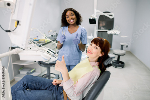 Healthy teeth and caries prevention concept. Young European woman at the dentist's chair during a dental examination and treatment, showing thumb up. Young African doctor dentist with dental tools