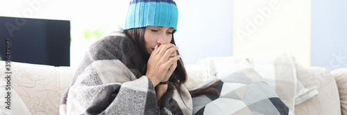 Sick woman in hat sit on sofa covered with blanket. Person hold hands together near his mouth and warm. photo