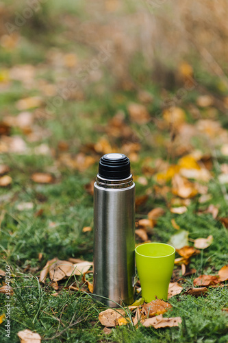 A metal thermos with tea and a green plastic cup stand on the grass with yellow maple leaves against the background of nature. Autumn portrait, advertising.