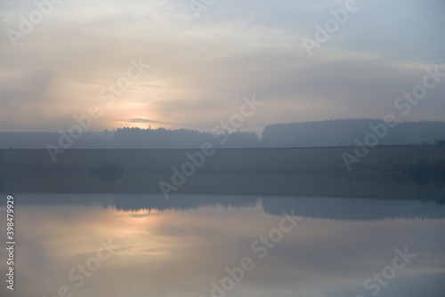 Pastel colored sunset behind trees over reservoir water. Hazy clouds