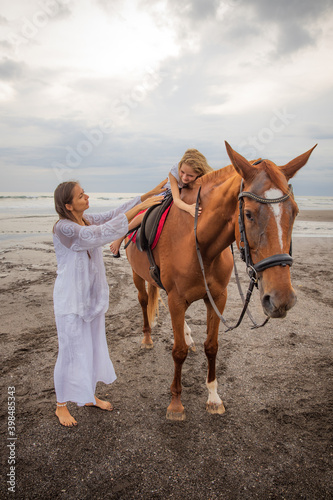 Cute little girl lays down on the withers of the horse. Mom and daughter spending time together. Mother's day. Copy space. Sunset time on the beach. Outdoor activities.