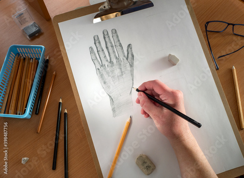 On the table are a drawing tablet with a sheet of paper, pencils, glasses and erasers. The human hand draws with a pencil on paper. Drawing training, lessons, hobbies. photo