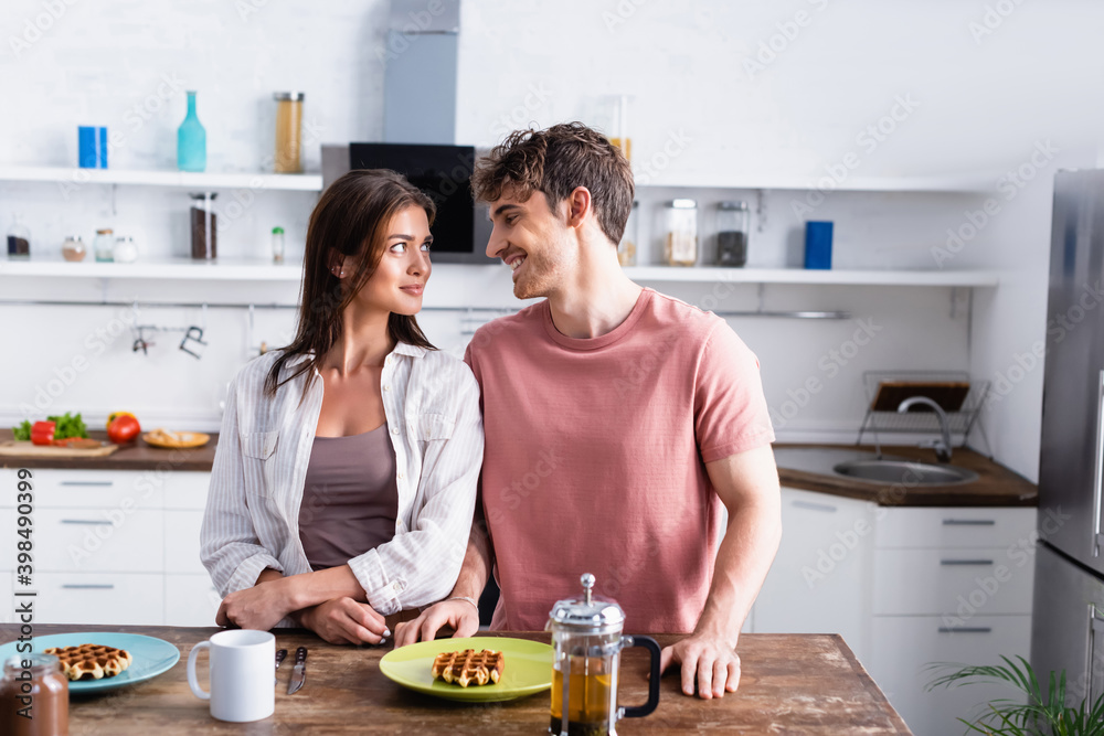 Smiling man standing near girlfriend, waffles and tea on kitchen table