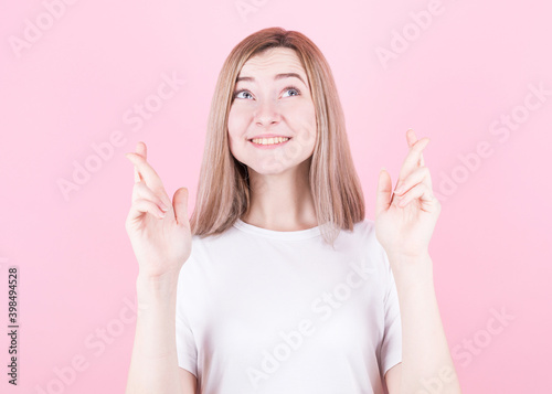Closeup of happy lovely young woman wears t shirt keeps fingers crossed and looks up, isolated over pink background