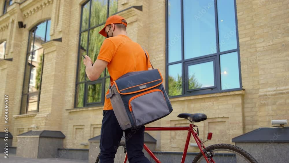 Masked courier using mobile phone outdoors. Uniform man leading bicycle street. Masked courier using mobile phone outdoors. Uniform man leading bicycle street.